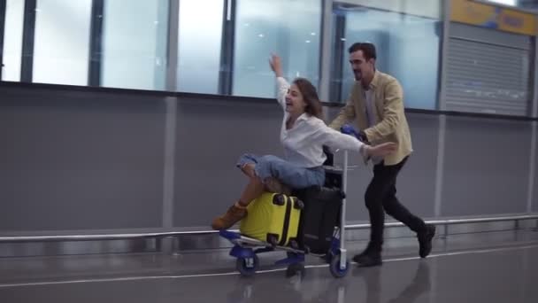 Lustiges Paar am Flughafen. Attraktive junge Frau und gutaussehender Mann mit Koffer sind bereit für die Reise. Spaß auf dem Gepäckwagen beim Warten auf die Abreise. Frau ahmt Fliegen nach, lächelt — Stockvideo
