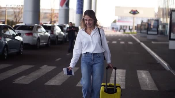 Opgewonden, stijlvolle vrouw lopen met trolley gele geval door lege luchthaven terminal buiten weg, slanke vrouwelijke dragen jeans en wit shirt. Gelukkige, lachende vrouw die loopt om haar vliegtuig te nemen met paspoort — Stockvideo