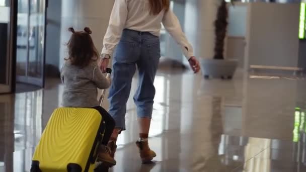 Luchthaven terminal, jonge onherkenbare moeder berijdt haar schattige dochter op een kleine gele koffer. Moeder met gele koffer en dochter hebben plezier voor hun vertrek — Stockvideo