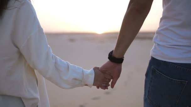 Mirato, riprese ravvicinate di una coppia che si tiene per mano mentre cammina in una relazione romantica sotto il sole e il cielo blu nel deserto. Due giovani amanti passeggiano sulla sabbia del deserto in abiti casual. Paesaggio sullo sfondo — Video Stock