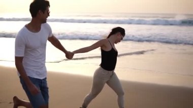 Güneş gözlüklü tatil çifti gün batımında romantik bir şekilde el ele tutuşuyorlar. Genç aşıklar plajda gündelik kıyafetlerle romantizmin tadını çıkarıyorlar.