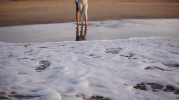 砂浜の海岸線に立つ若い夫婦は、海からの夕日の波を見る。サングラスとカジュアルな服を着て。泡の波が来る。正面図 — ストック動画