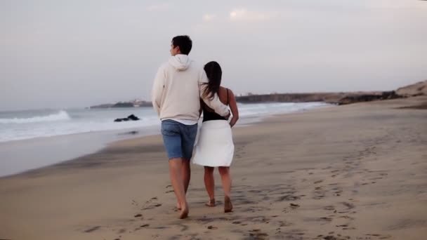 Relaxou jovem casal na praia andando no homem do amor segurando sua menina por trás. Mulher e homem relaxante em férias de viagem, homem vestindo roupas casuais, mulher toalha branca na barriga. Devagar. — Vídeo de Stock