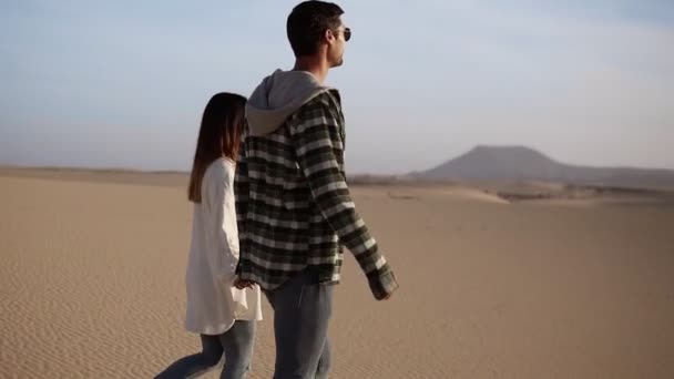 幸福的夫妻手牵着手，在阳光下漫步，在蓝天的沙漠中漫步。两个年轻的恋人穿着休闲装在沙滩上散步，面带微笑 — 图库视频影像