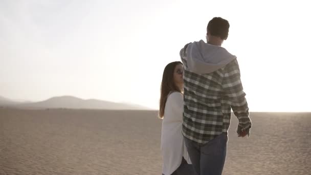 Genç çift çölde yürüyor. Uzun boylu genç bir adam batan güneşe karşı güzel bir esmer kızı öpüyor. Çöl, kumlu sahil, tatil ve dinlenme. Balayı konsepti — Stok video