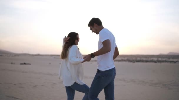 カジュアルな白いTシャツを着た素敵な陽気な笑いのカップル。暗い髪の男は彼の女性と回転し、移動中の手を握っている。空の砂漠で自然の中で踊る — ストック動画