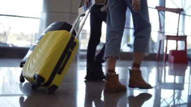 Bir çift gezgin bavullarını taşıyorlar ve terminalin arka manzaralı salonunda ilerliyorlar. İnsanlar binmeye ve gitmeye hazırlanıyor. Yuvarlanan bavulları kapat.