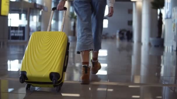 Elegante mujer caminar con el caso de carro amarillo por el aeropuerto vacío, vista media baja de las piernas delgadas hembra y bolsa de ruedas medianas, tiro de seguimiento suave. Usando jeans y botas marrones. Vista rara — Vídeos de Stock