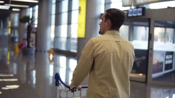 Brunett, skäggig man i beige skjorta går med bagagevagn på den internationella flygplatsen. Går i brådska, sällsynta spårningsbilder — Stockvideo