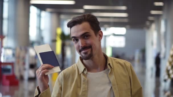 Mutlu, yakışıklı, sakallı Amerikalı adam pasaportunu uçak biletleriyle gösterip el sallıyor. Havaalanında dikilirken kameraya bakıp gülümsüyor. — Stok video