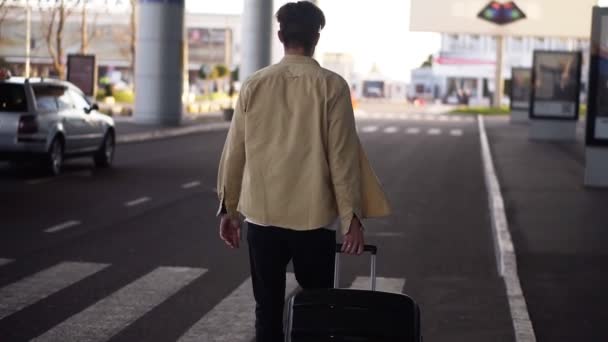 Уверенный, высокий мужчина в повседневной жизни с чёрным багажом, идущий до терминала аэропорта по дороге. Медленное движение. Редкий вид. Размытый задний план — стоковое видео