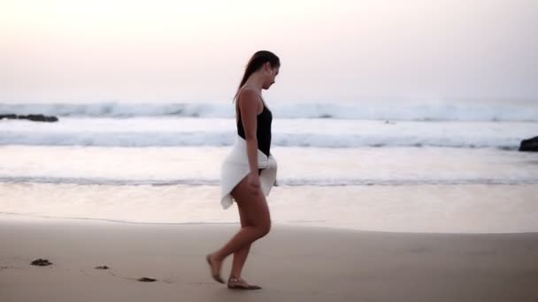 Menina sexy em um maiô preto e camisa branca apertado na barriga caminha sobre uma praia de areia no oceano - linda menina caucasiana de cabelos longos caminha sozinha à beira-mar em humor despreocupado, correndo. Vista lateral — Vídeo de Stock