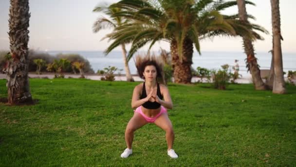 Kaukasische Frau macht Sport im tropischen Park. Kniebeugen auf dem Gras der Beine Muskeln intensiv springen nach oben. In sexy, rosa Hemden und BH. Ozean, Bäume im Hintergrund — Stockvideo