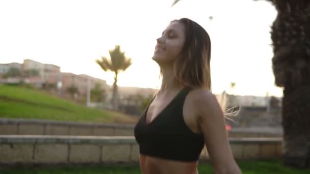 Retrato de una joven chica positiva haciendo ejercicio al aire libre en el parque tropical. Chica sonriente en sujetador negro saltando, calentándose como parte del entrenamiento. Movimiento lento — Vídeo de stock