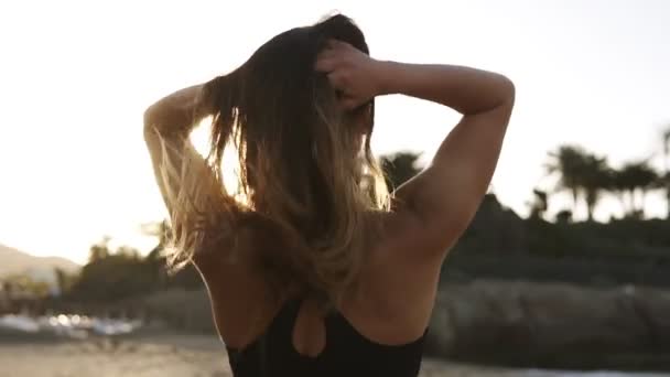 Zabawna młoda kobieta ciesząca się wakacjami na letniej plaży. Zmysłowa kobieta z wietrznymi włosami spaceruje po plaży w czarnym staniku nad morzem. Patrząc wstecz od ramienia — Wideo stockowe