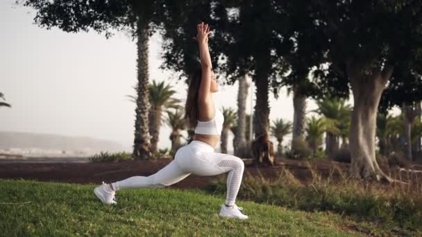 Sportvrouw in witte sportkleding die yoga poseert of zich buiten uitstrekt. Een atletisch meisje dat zich uitstrekt in het park. Fitness vrouw doet yoga oefeningen op groen gras in slow motion. Palmbomen op — Stockvideo