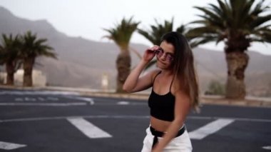 Süper pozitif, heyecanlı, beyaz pantolonlu, yolda dans eden şık bir kadın. Arka planda palmiye ağaçları ve diğer tropikal bitkiler vardır. Dönen, dans eden, nefes alan, mutlu ruh hali