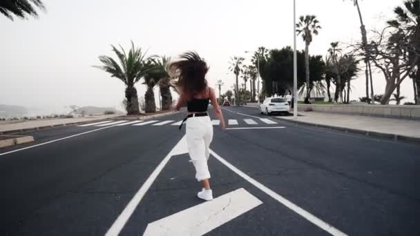 Emocionado joven mujer con estilo en pantalones blancos posando, bailando en la carretera. En el fondo hay palmeras y otra vegetación tropical. Girando, bailando, respirando, de buen humor, oliendo. Despacio. — Vídeo de stock