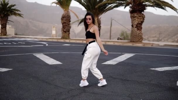 Stylish young woman in sunglasses, black top and white pants dances funky in the middle of the road - slow motion - Woman freerly moves her body. Palm trees and hills on background — Stock Video