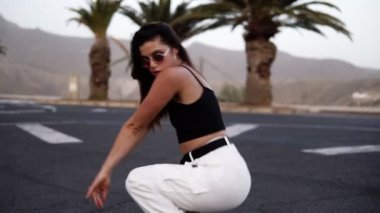 Beyazlı seksi kız, arka planda tropik ağaçlar ve tepelerle yolda dans ediyor. Seksi uzun saçlı esmer kadın dans alıştırması yapıyor, vücudunu profesyonel olarak hareket ettiriyor. Yavaş çekim