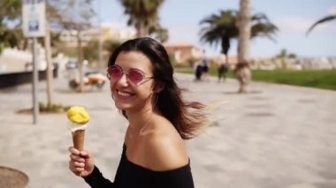 Güneş gözlüklü güzel bir kadın parkta palmiye ağaçlarıyla dondurma yiyor. Yaklaş. Ağır çekim. Lens fişeği. Genç ve çekici bir kadın, açık havada, yeşil güneşli yaz mevsiminde dondurmanın tadını çıkarıyor.