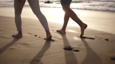 Sahilde çıplak ayakla yürüyen rahat bir çift. Birlikte vakit geçiriyorlar, ıslak kumda ayak izleri bırakıyorlar, el ele tutuşuyorlar. Arka planda gün batımı var. Nadir görünüm
