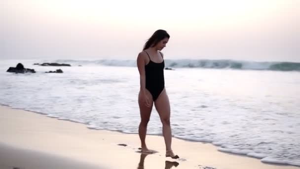 Sexy girl in einem schwarzen Badeanzug geht über einen Sandstrand am Meer - wunderschöne langhaarige kaukasische Mädchen geht allein am Meer in unbeschwerter Stimmung, läuft, plantscht das Wasser. Milder Sonnenuntergang auf der — Stockvideo