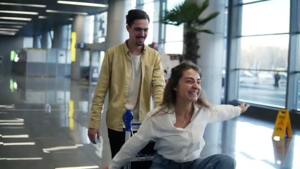 Pareja romántica en el aeropuerto. Atractiva joven mujer y hombre guapo con maletas están listos para viajar. Diviértete en el carrito de equipaje mientras esperas la salida. Mujer imitando volar, oler — Vídeos de Stock