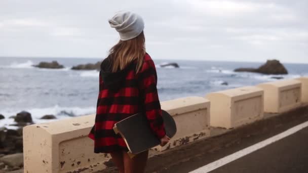 Zeldzaam uitzicht van sportieve gebouwde vrouw in hoed, jas en witte sneakers genieten van de tijd aan de kust op een bewolkte dag bij zonsondergang, wandelen, het houden van een skateboard, slow motion — Stockvideo