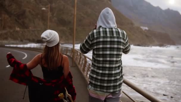 İki genç insan, deniz kıyısının arka planına karşı otoyol boyunca yürüyorlar. Seyahat ve özgürlük, maceralar ve seyahat için yol tarifi. Kızın elinde kaykay var. Nadir görünüm — Stok video