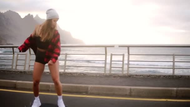 Sport, stile di vita, estremi e concetto di persone Bella ragazza a cavallo longboard dalla strada costiera in tempo nuvoloso. Ritratto hipster ragazza sorridente con un longboard al tramonto. Al rallentatore. Vista laterale — Video Stock