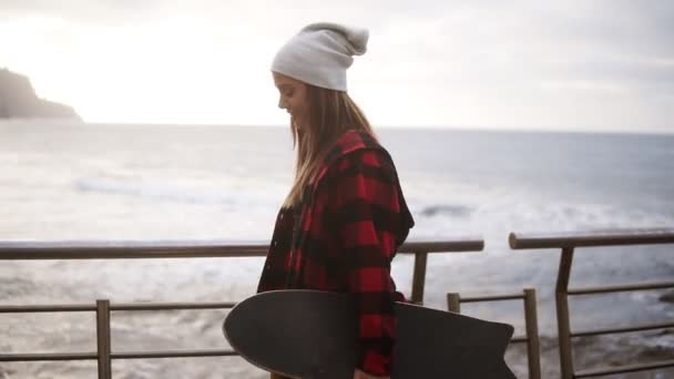 帽子の中のスポーティな女性の映像を閉じ、日没の曇りの日に海辺で時間を楽しんでコートをプレイし、自由に歩き、スケートボードを保持します。風が彼女の髪をなびかせ、ポーズをとり、カメラを微笑んだ — ストック動画