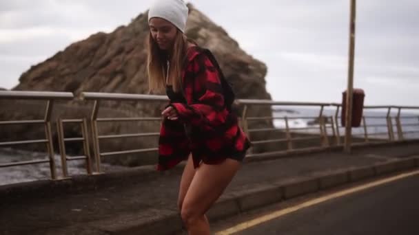 Jeune jolie belle brune hipster femme en manteau à carreaux s'amusant à monter skateboard longboard descente sur belle route au ralenti. Côte, temps nuageux — Video