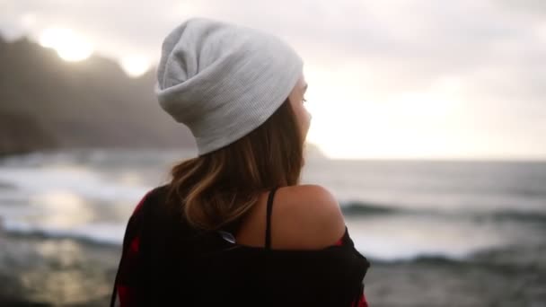 A nő az óceánt nézi - egy barna nő hátulról néz ki az óceánra a hegyek előtt, majd oldalra fordítja a fejét mosolyogva a válla fölött. Homályos háttér — Stock videók