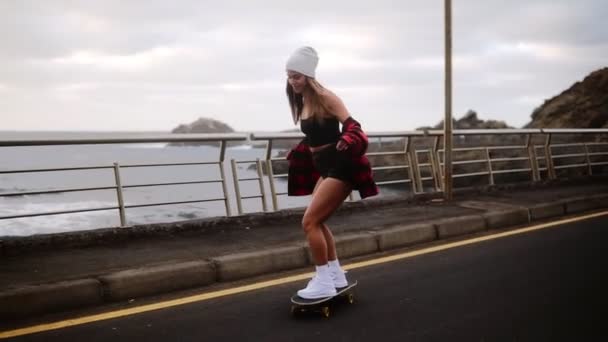 Ung, bekymringsløs, vakker brunette hipster kvinne i rutete frakk som henger på hender og har det gøy med å kjøre skateboard longboard på en vakker vei i sakte film. Kystlinje, tåkete vær – stockvideo