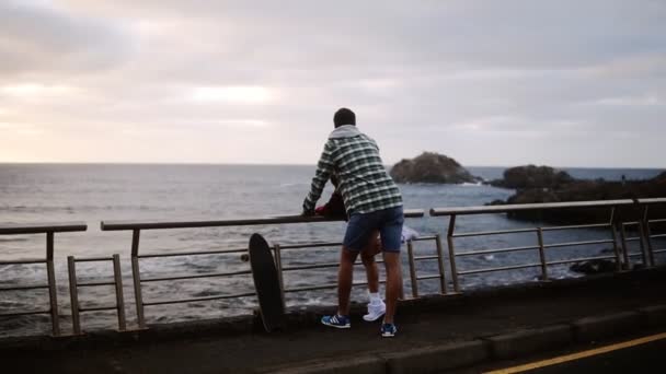 Woman and standing together, looking on ocean - Young couple shown from the side look out over an ocean in front of the hills, talking, embracing. Cloudy weather. Loving couple — Stock Video