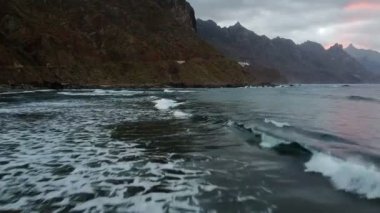 Okyanus ve dağlar manzaralı güzel bir sahil. Sabah alacakaranlığı, alçak hava manzarası köpüklü sahil dalgaları geliyor. Arka planda kahverengi yüksek tepeler