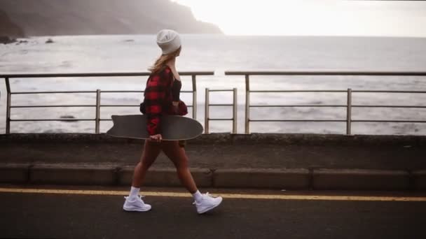 Sidovy över muskulös, sportig kvinna i hatt, rock och shorts njuter av tid vid havet på en molnig dag vid solnedgången, promenader freerly, hålla en skateboard. Vind fladdrar hennes hår. Stormigt hav på — Stockvideo