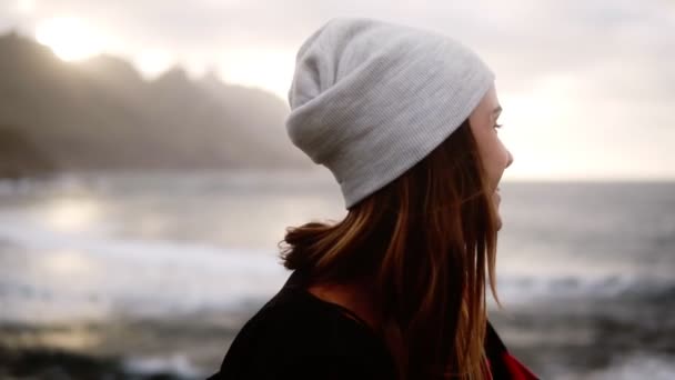 Portrét krásné ženy v klobouku, červený kostkovaný kabát, jak si užívá času u moře za zamračeného dne, jak stojí a opírá se o zábradlí skateboardem. Oceán z promenády, zpomalený pohyb — Stock video