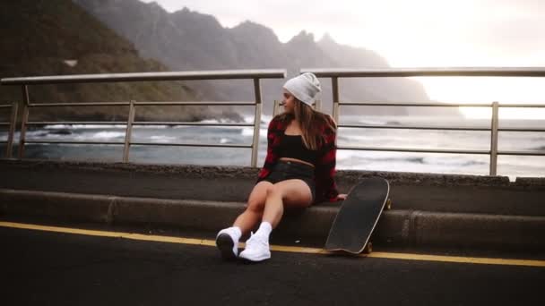 Dívka sedí s longboardem na hranici na pobřeží asfaltové silnici na pozadí kopce a mlhavý oceán. Uvolňující přestávka. Zataženo — Stock video