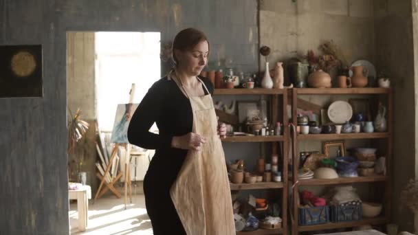 Filmati al rallentatore di giovane donna caucasica in abiti casual in piedi in laboratorio di ceramica, con ceramiche e forniture su scaffali e tavolo con strumenti in mezzo, e mettere su grembiule beige — Video Stock