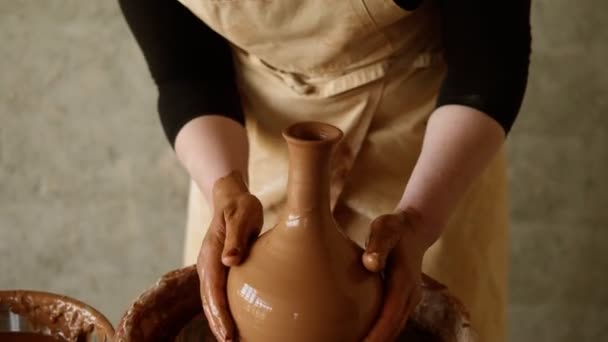 Vrolijke, jonge, goed uitziende vrouw met een schort in aardewerk die met handen kleikruik maakt op pottenbakkerswiel. Vrouw glimlachend naar een camera — Stockvideo