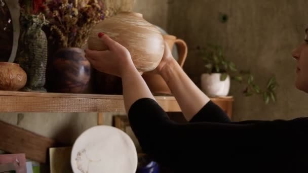 Boční pohled portrét současné hrnčířky, jak dává ručně vyrobenou vázu na polici ve studiu, spokojená se svou prací, usmívá se do kamery. Zpomalený pohyb — Stock video