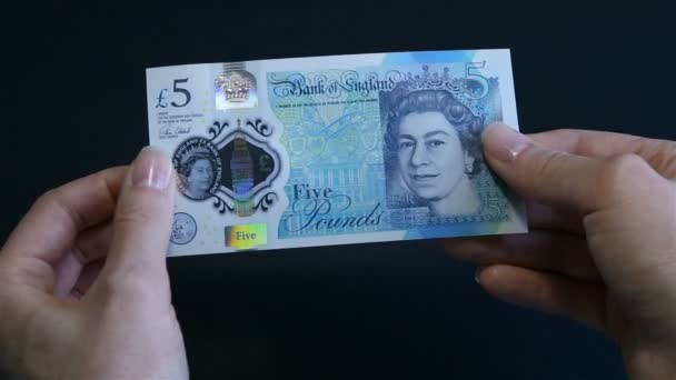Женщины держат в руках и показывают новую банкноту весом 5 фунтов, защитные знаки британской валюты. Проверяю деньги на подлинность. Фунт стерлингов крупным планом. Чёрный фон . — стоковое видео