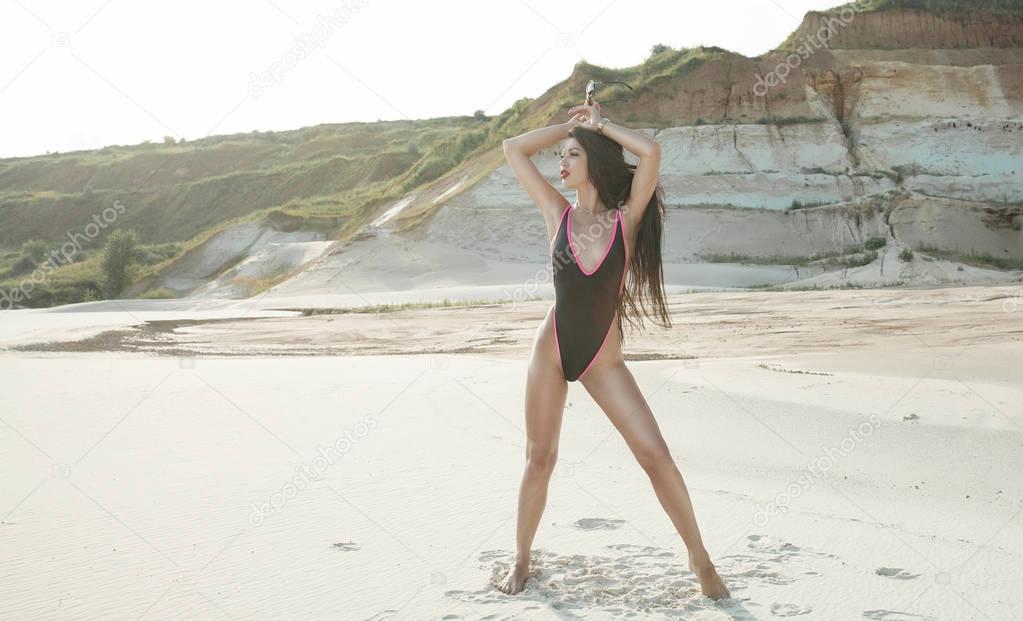 Sexy brunette Woman in black swimsuit with long black hair and red lips on the background of sands career