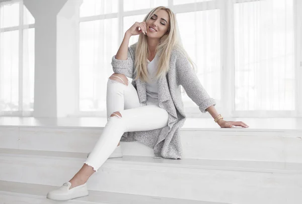 Retrato de una atractiva mujer rubia joven de moda en jeans blancos y abrigo gris . — Foto de Stock