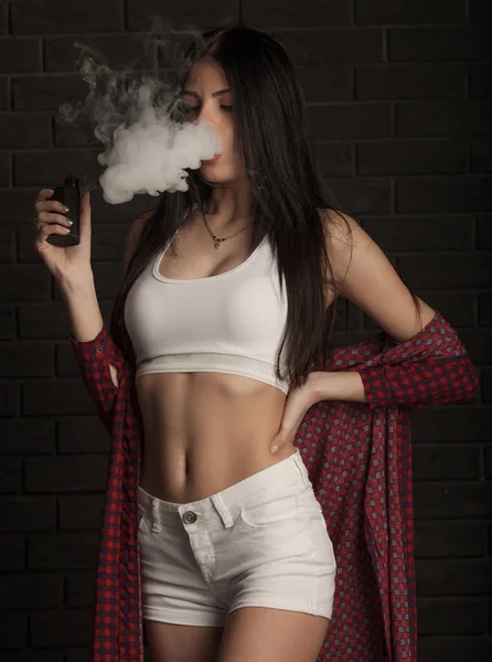 Jovem mulher bonita morena fumando, fumando e-cigarro com fumaça no fundo preto . — Fotografia de Stock