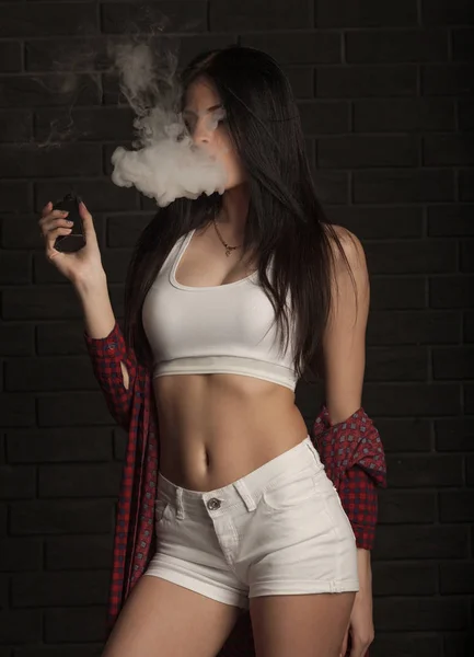 Jovem mulher bonita morena fumando, fumando e-cigarro com fumaça no fundo preto . — Fotografia de Stock