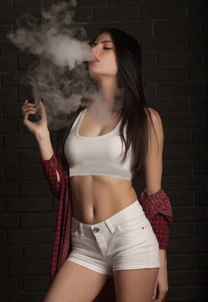 Young beautiful brunette woman smoking, vaping e-cigarette with smoke on black background. — Stock Photo, Image