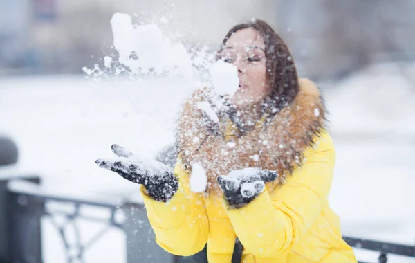 Happy beautiful young woman blowing snowflakes from her hands in a winter day — Stock Photo, Image